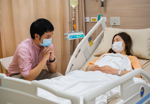 Picture of a female patient sleeping in a swing bed and a male visitor sitting next to her bedside. They are both wearing mask.